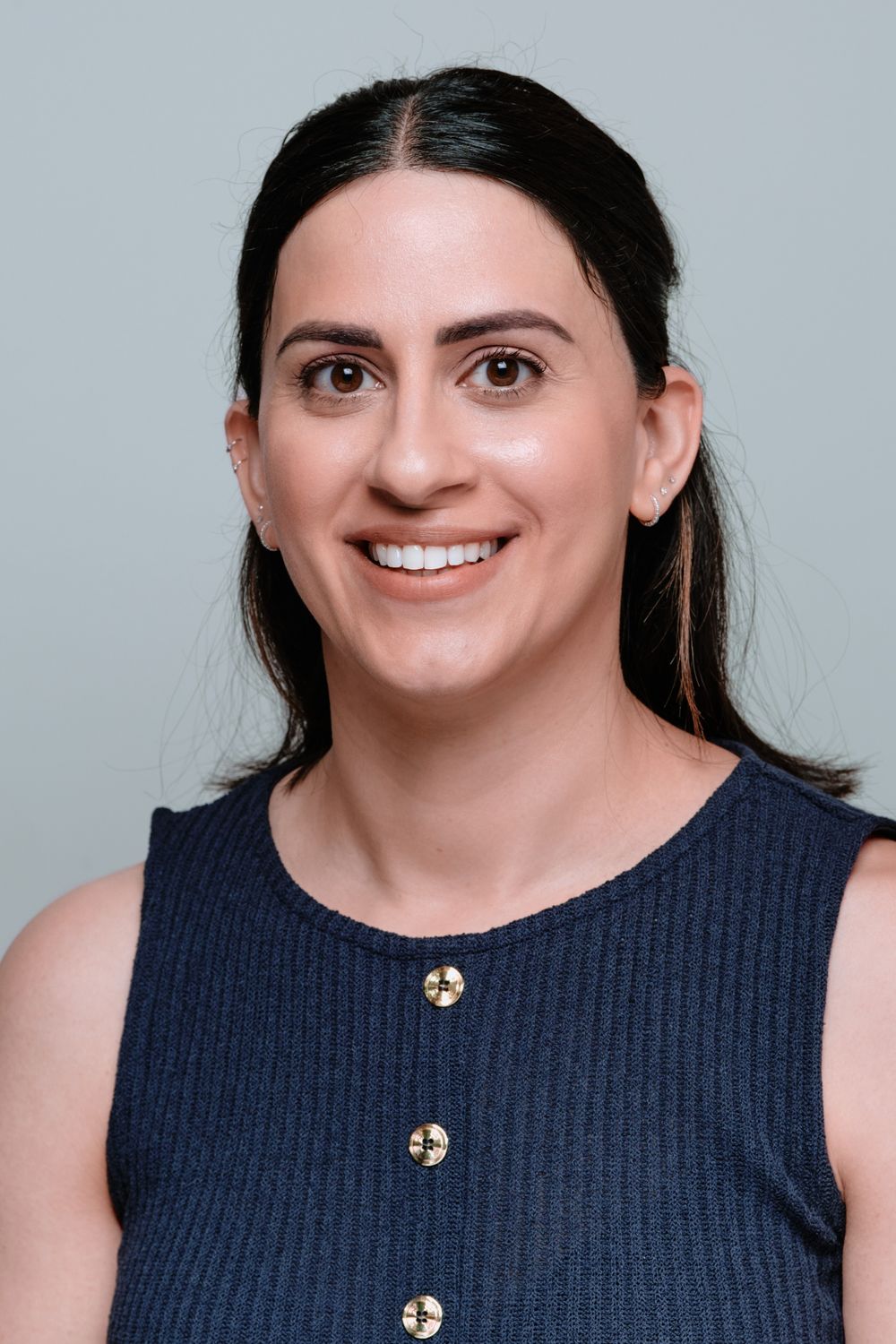 Professional headshot of someone wearing a navy sleeveless top with gold buttons against a light gray background.