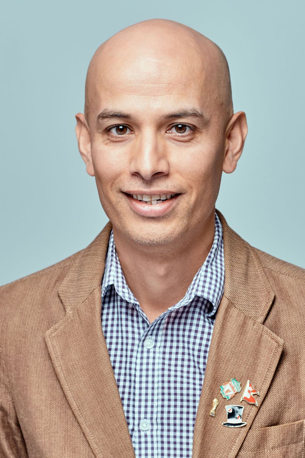 Professional headshot of a bald man wearing a brown blazer and blue gingham check shirt against a light blue background.
