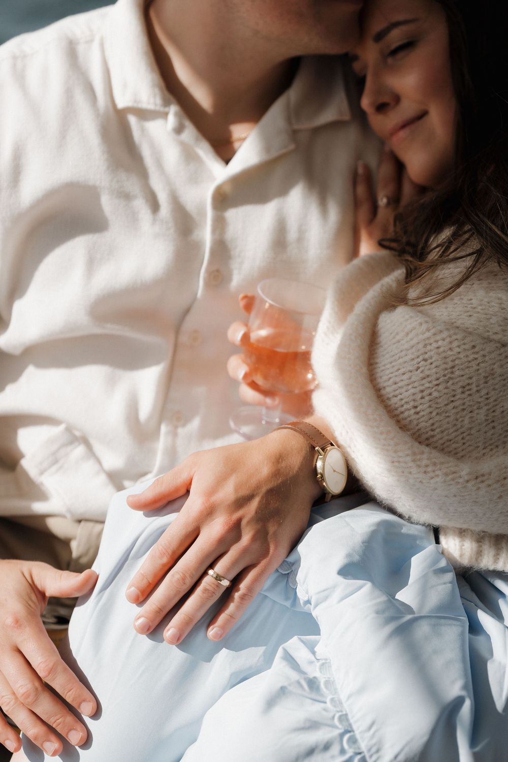Close-up of a couple embracing on a boat, a man wearing a watch and ring while the woman holds a glass of rose wine.