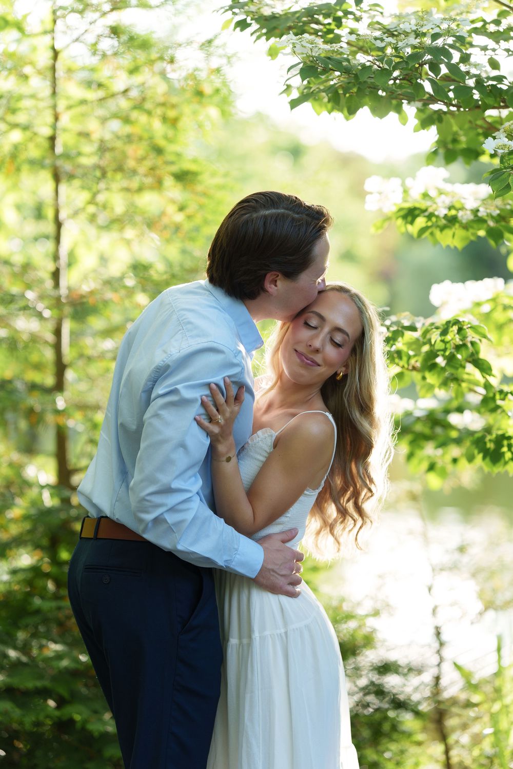 Groom kissing bride on cheek as she smiles serenely in a sunlit green forest setting.