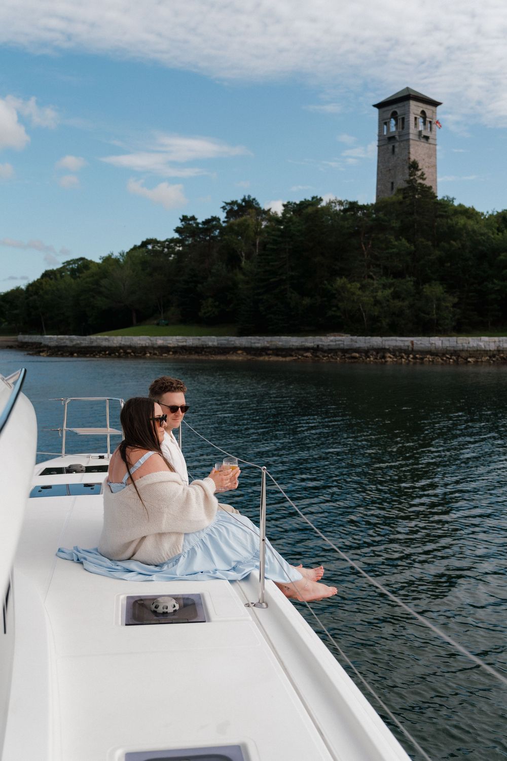 Couple sitting on yacht bow with wine, historic Dingle tower visible on shore.