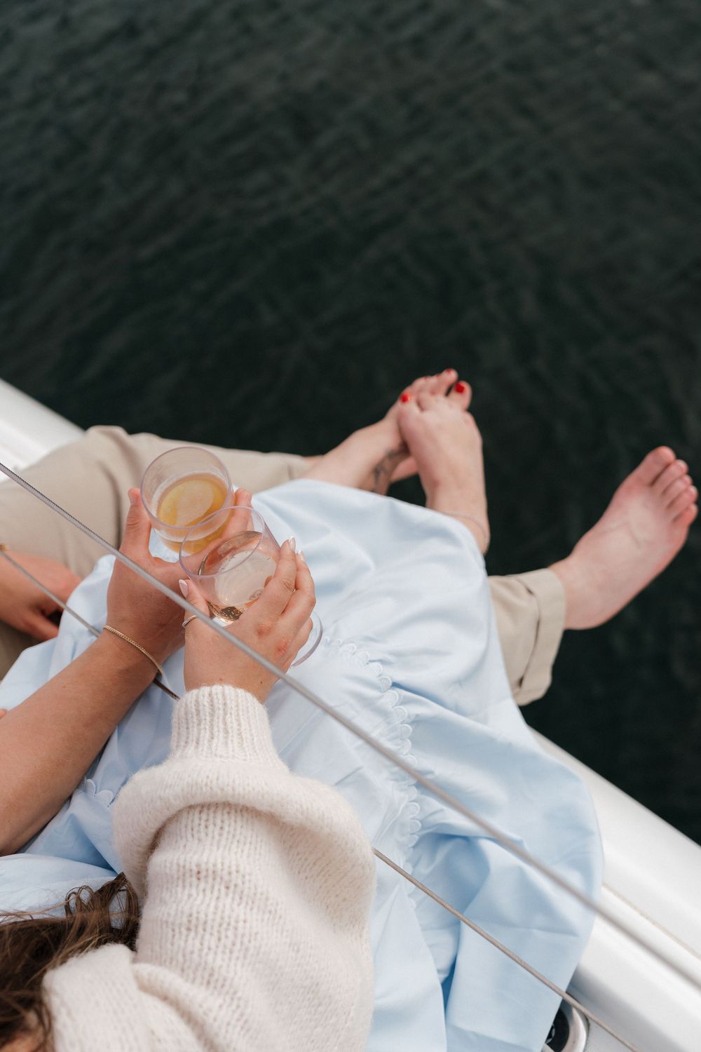 Couple relaxing on a sailboat deck, woman in blue dress holding a cocktail with bare feet dangling over the water.