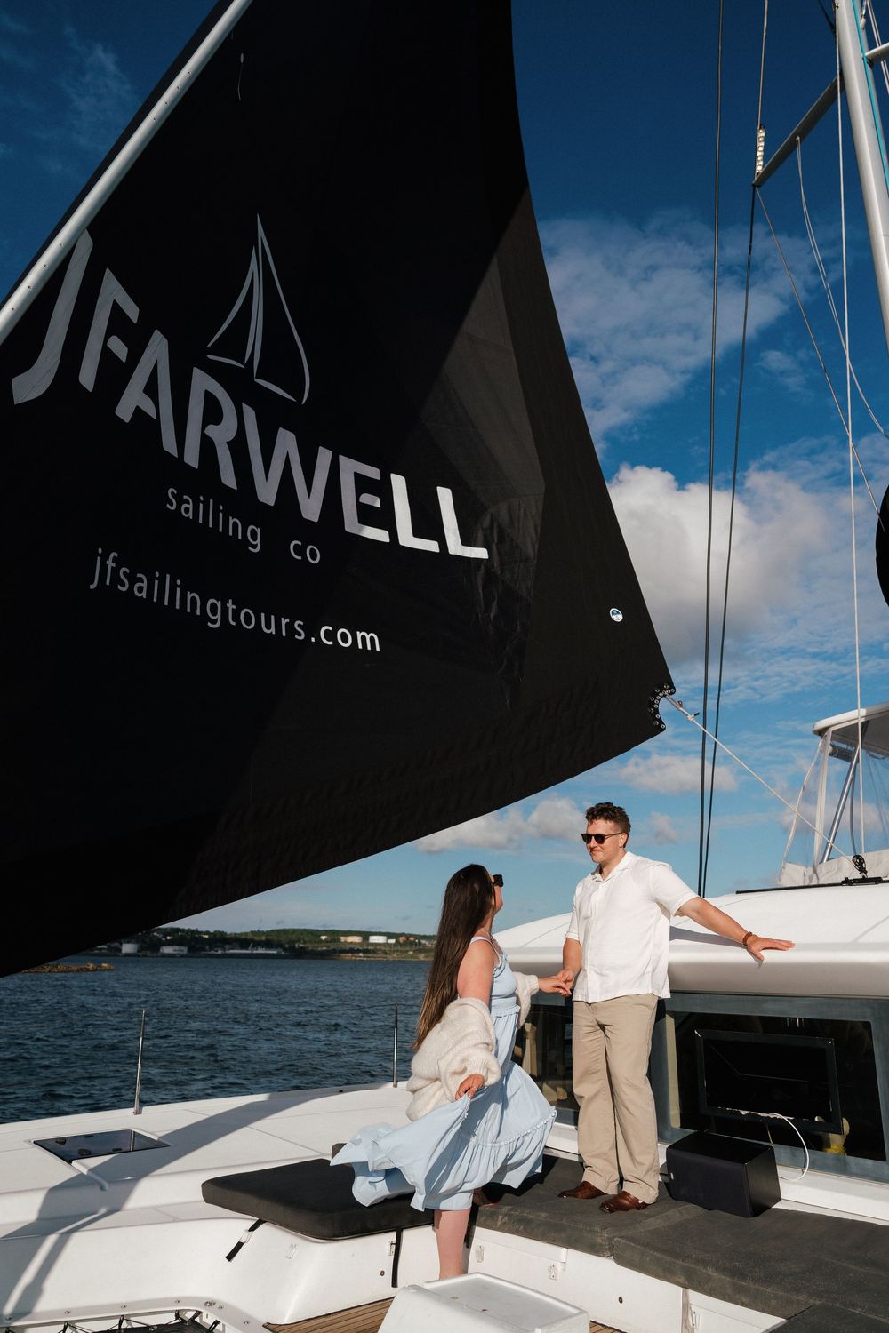 Couple standing on a sailboat deck beneath a JFarwell Sailing Co. black sail on a sunny day.