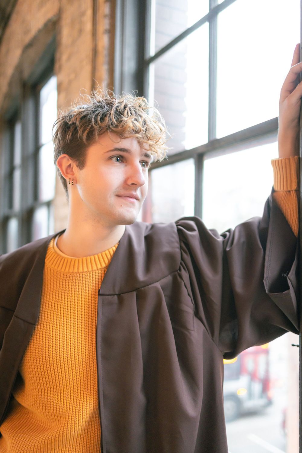 Young man in orange sweater and brown coat gazes out window in brick building.