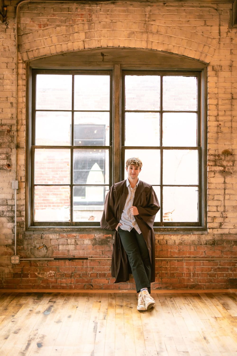 Young man in long brown coat leans against large window in rustic brick loft.