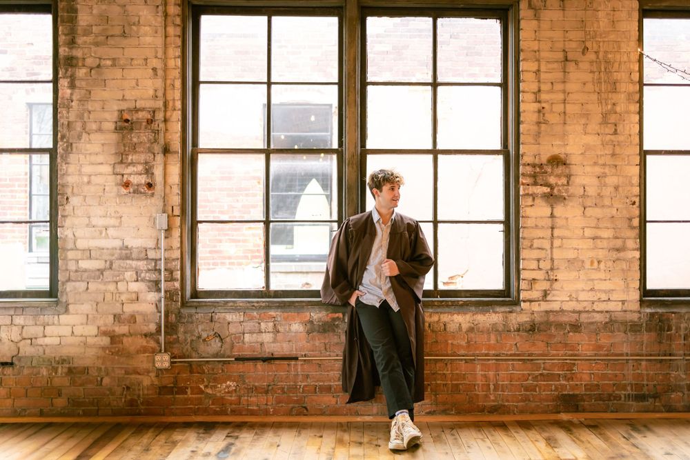 Young male graduate in dark academic gown standing confidently in a rustic brick building with large windows.