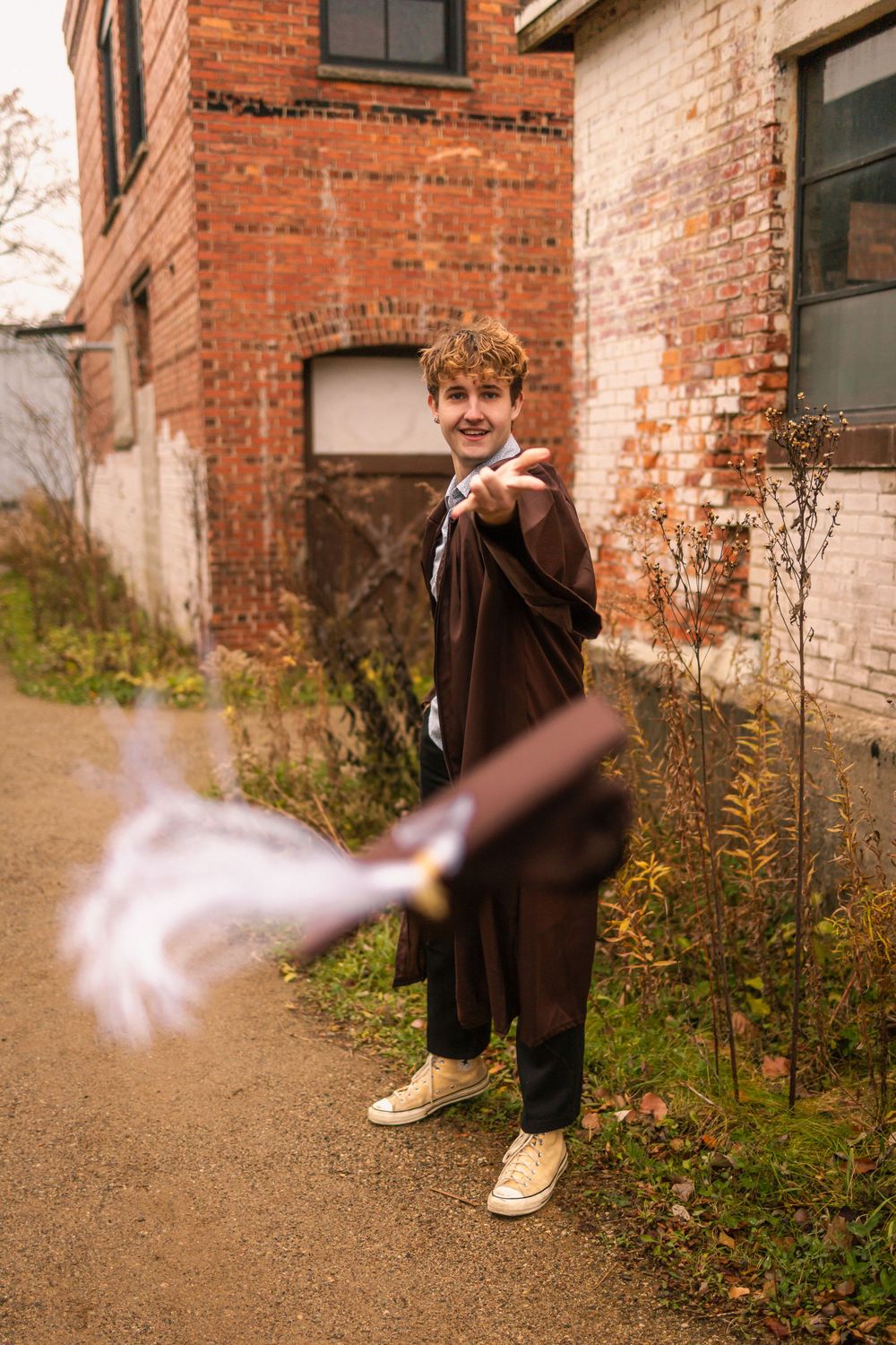 Smiling male graduate in brown gown tossing his graduation cap outdoors near a rustic brick building.