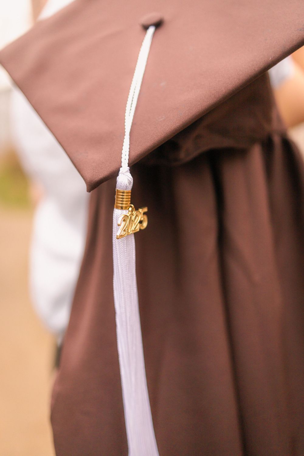 Close-up of brown graduation cap and gown with white tassel and gold 2025 charm.