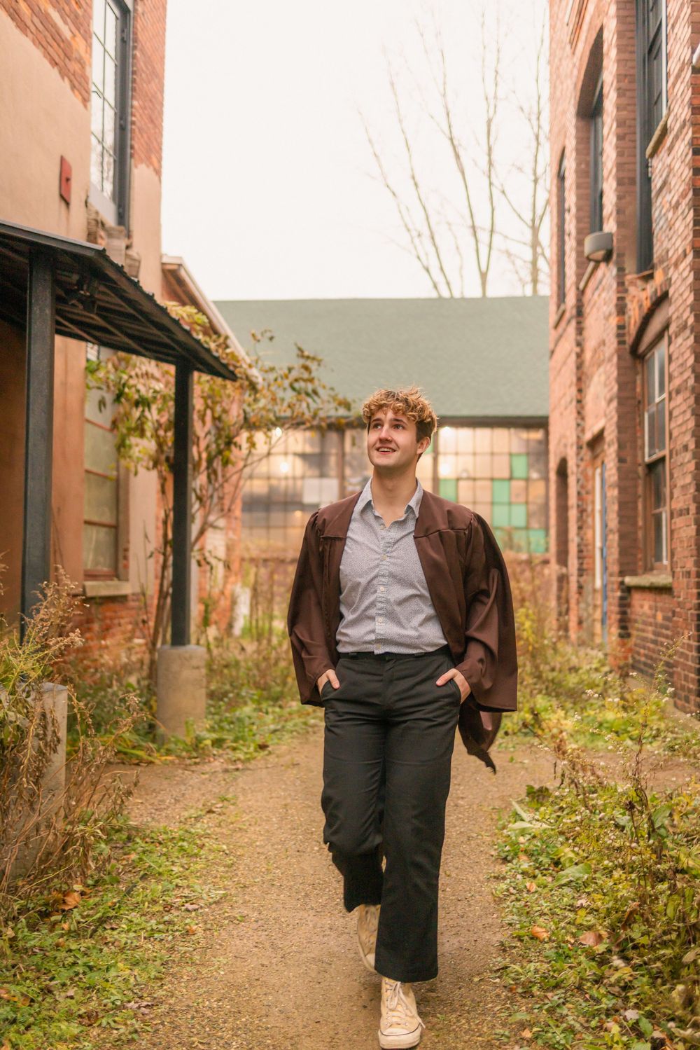 Young man with curly red hair walks confidently between brick buildings wearing dark trousers and a brown jacket.