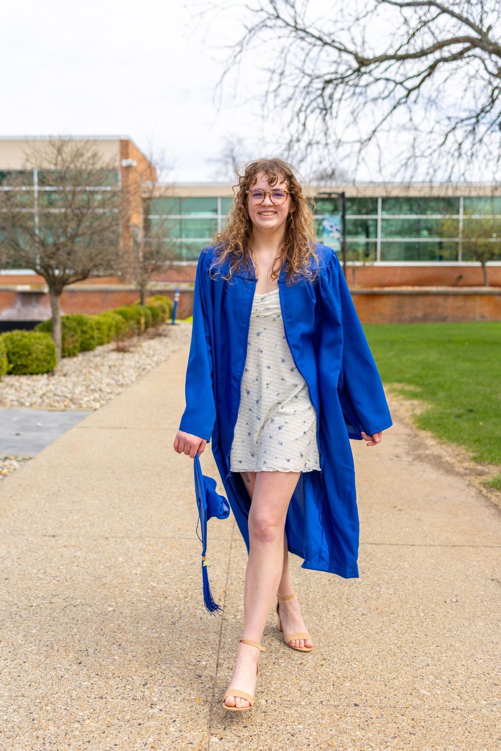 Young woman in blue graduation gown and cap walks on campus pathway, smiling, wearing glasses and floral dress.