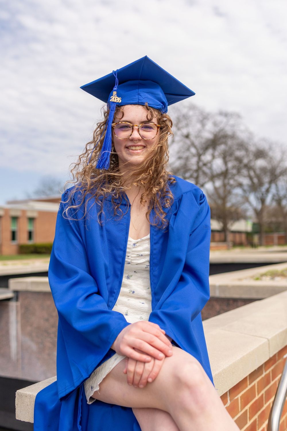Smiling young woman in blue graduation cap and gown sitting outdoors, celebrating her academic achievement.