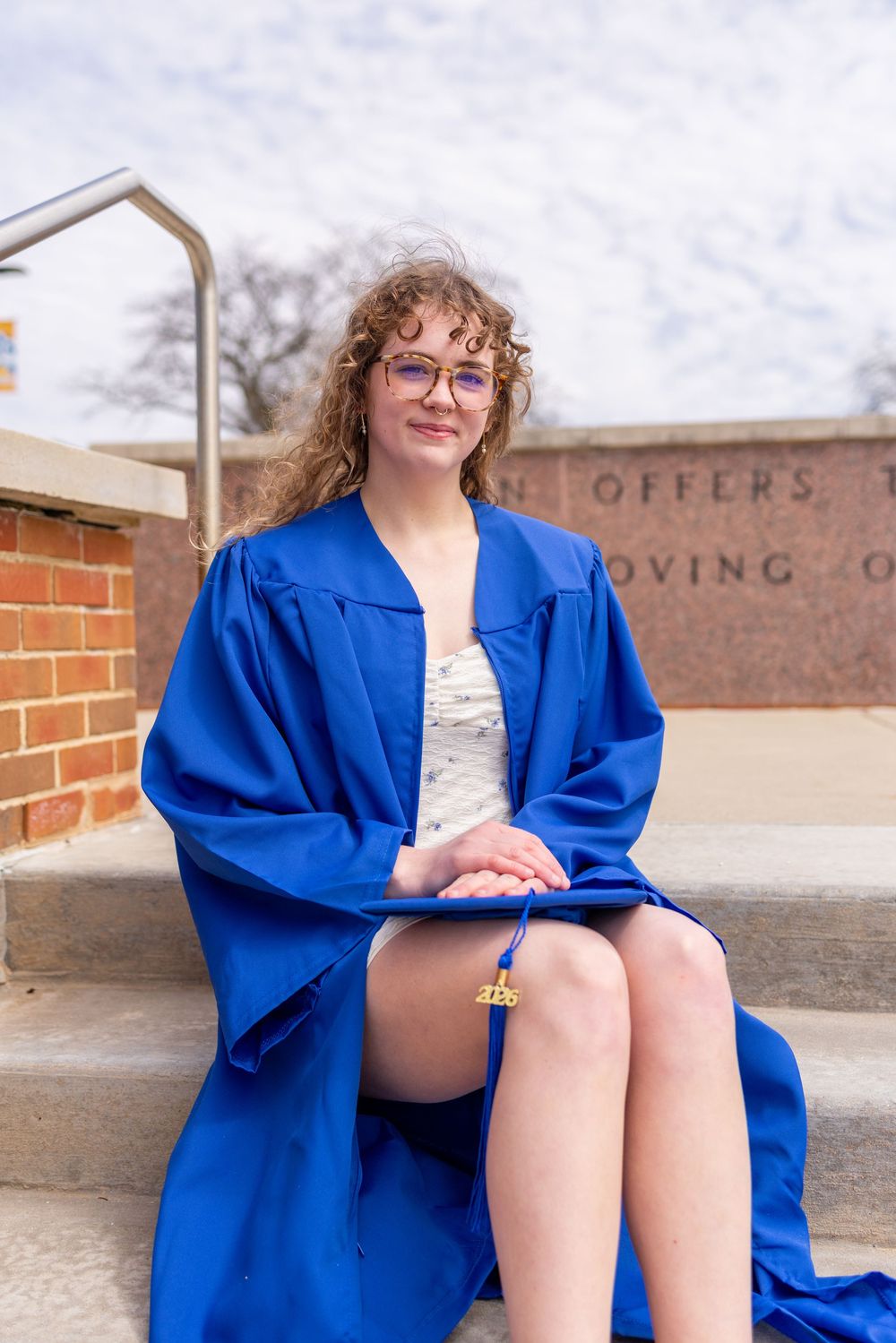 Young woman in blue graduation gown and glasses sits on steps holding diploma, smiling outdoors.