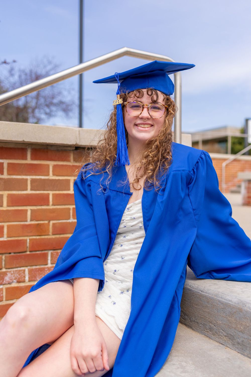 Young woman in blue graduation cap and gown sitting on steps, smiling, wearing glasses outdoors.