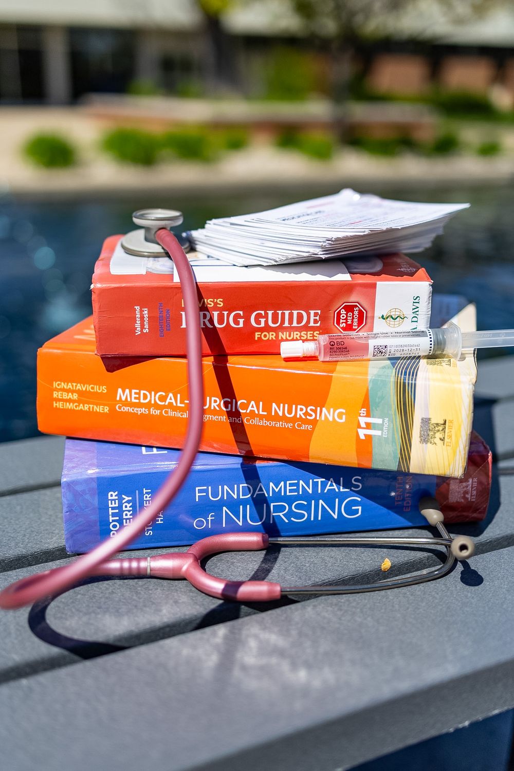 Stack of nursing textbooks including Fundamentals of Nursing and Drug Guide with a stethoscope outdoors.