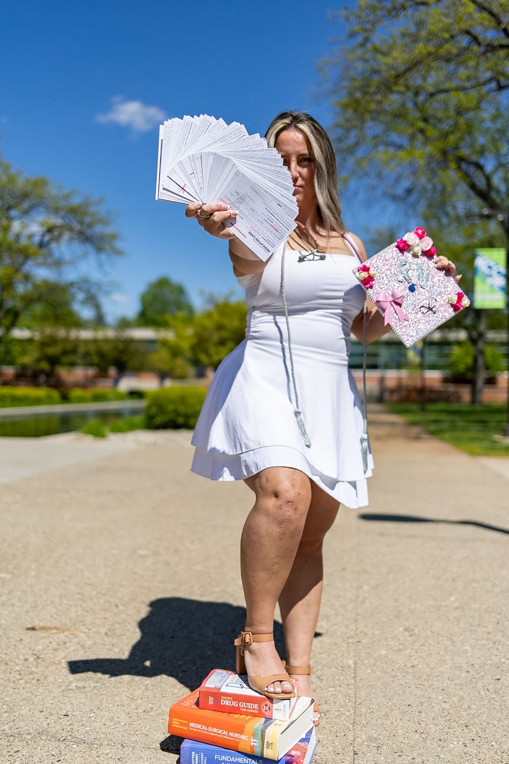 Woman in white dress holding papers and decorated cap, standing on books outdoors on a sunny day.