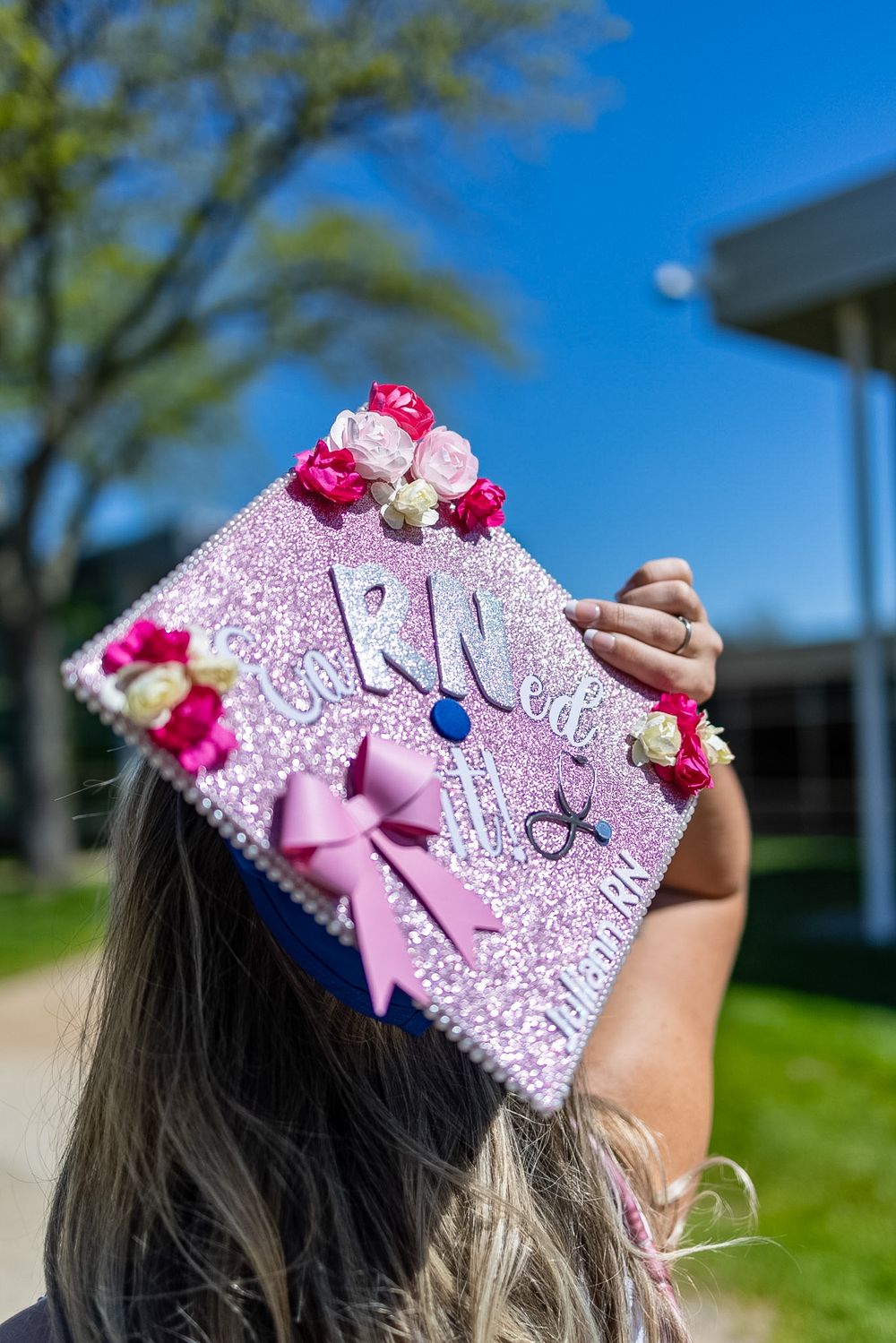 Decorated pink glitter RN nursing graduation cap with flowers and bow held up against blue sky.