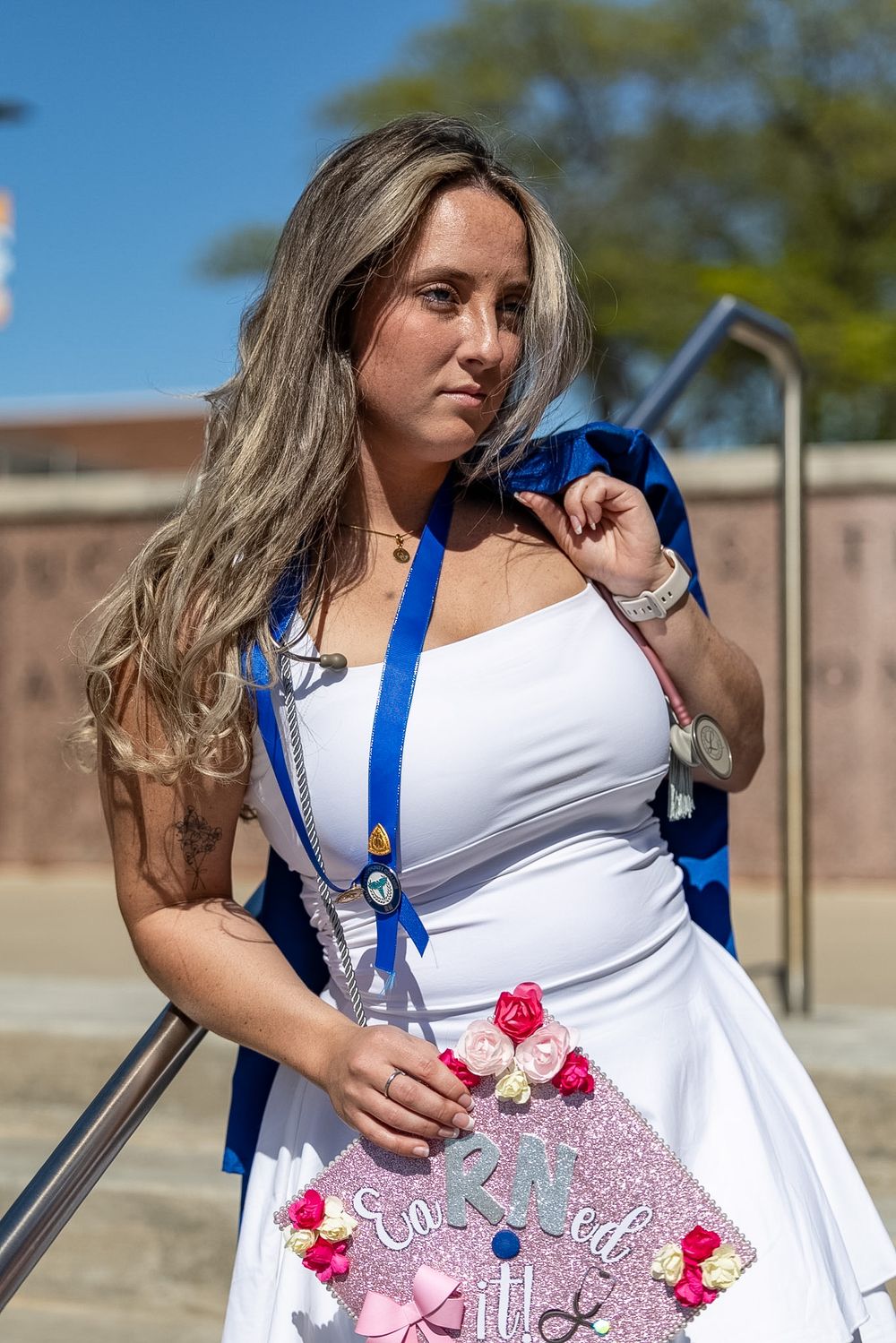 Graduate in white dress holding decorated cap with blue sash on sunny day near brick building.