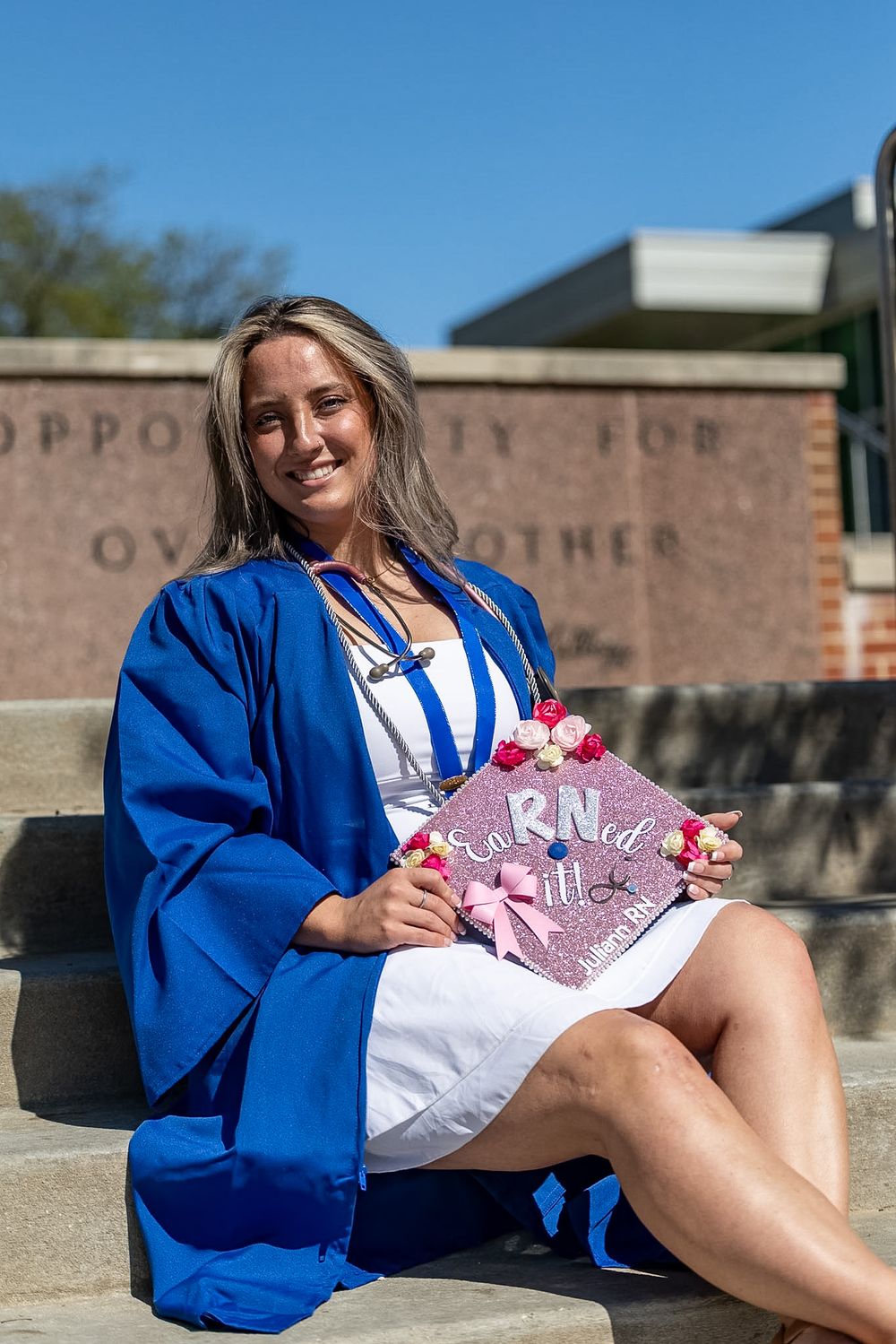 Smiling nursing graduate in blue gown sits on steps holding decorated pink graduation cap outdoors.