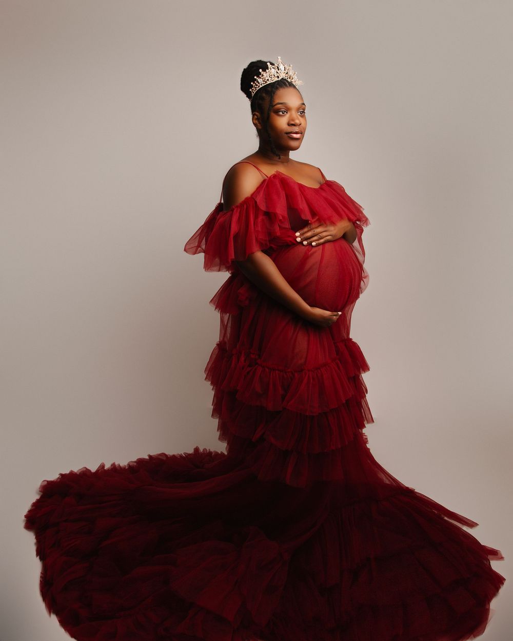 Regal maternity portrait in layered red tulle gown and crown against neutral backdrop.