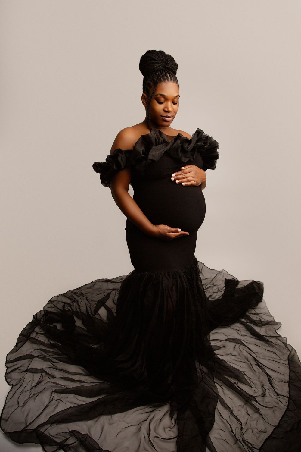 Elegant maternity portrait in flowing black off-shoulder gown against neutral background.