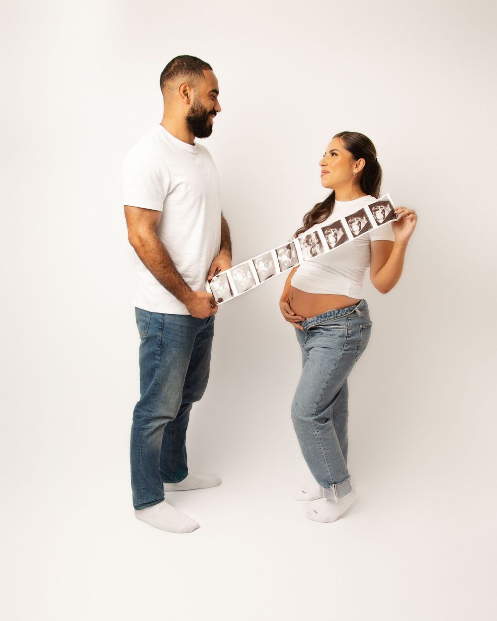 Expecting couple holding ultrasound strip in casual jeans and white shirts.