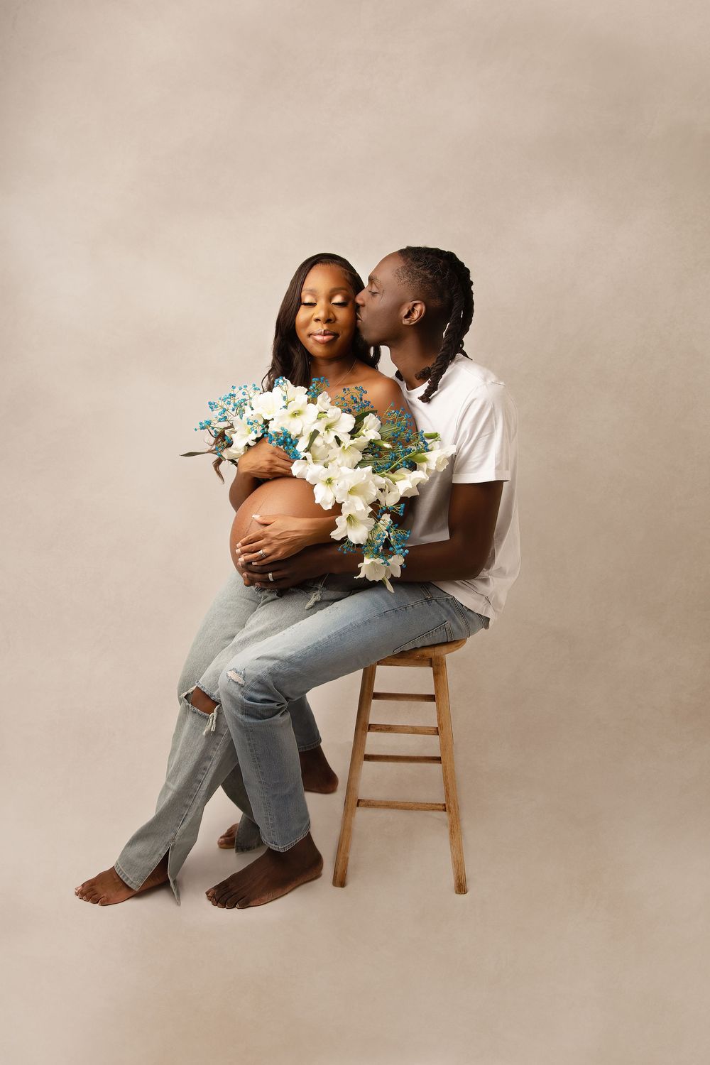 Romantic maternity photo with couple holding white flowers against beige backdrop.