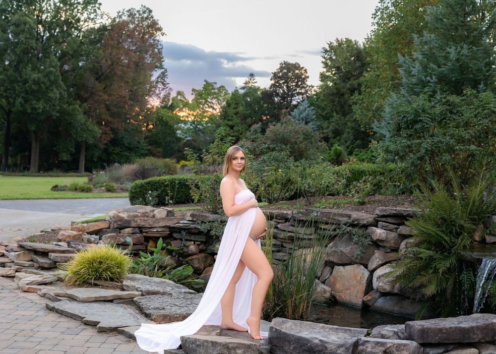 A white flowing dress contrasts against a stone wall and lush garden landscape at sunset at a maternity session