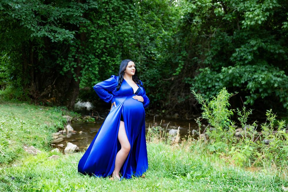 A maternity session in a flowing royal blue dress poses gracefully in a lush green garden setting outdoors.