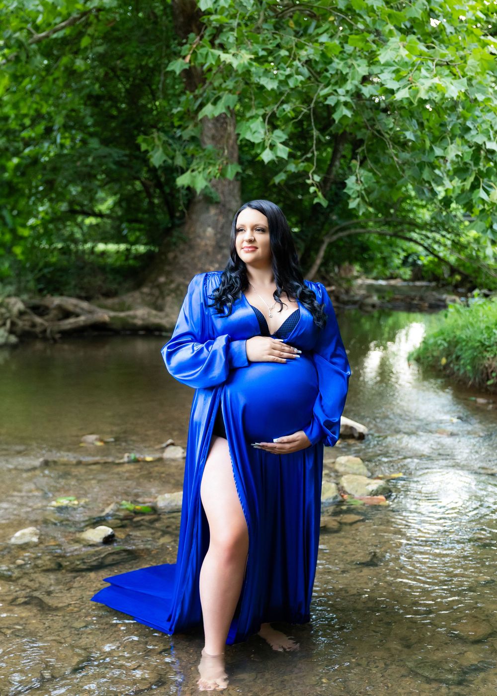 A maternity session in a royal blue dress poses gracefully by a rocky stream in a lush forest setting.