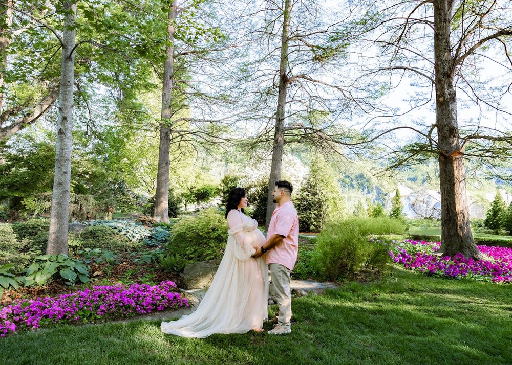 Couple in wedding attire sharing a romantic moment in a lush garden with tall trees and vibrant pink flowers.