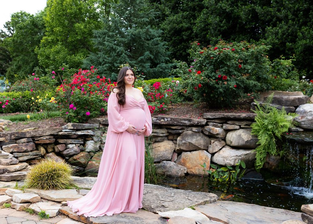In a garden setting with red roses and a stone wall waterfall, someone poses in a flowing pink maternity gown.