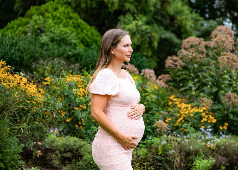 Pregnant woman in pink lace dress cradling baby bump in lush garden with yellow and green flowers.