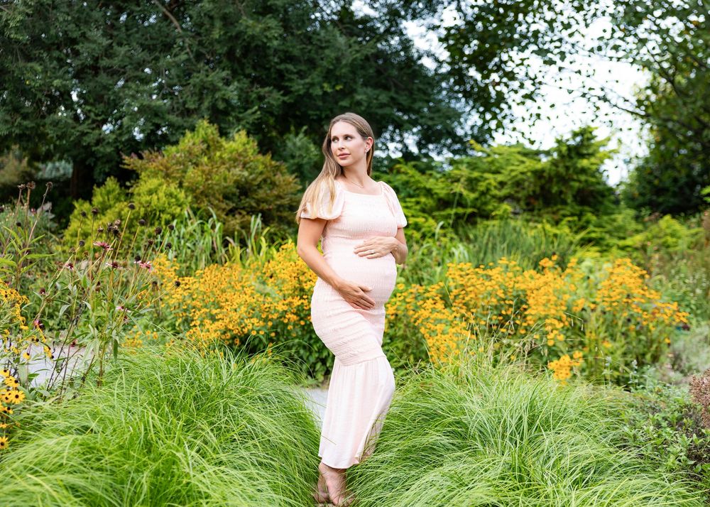 Pregnant woman in pink dress posing in lush garden with yellow flowers during maternity photo shoot.