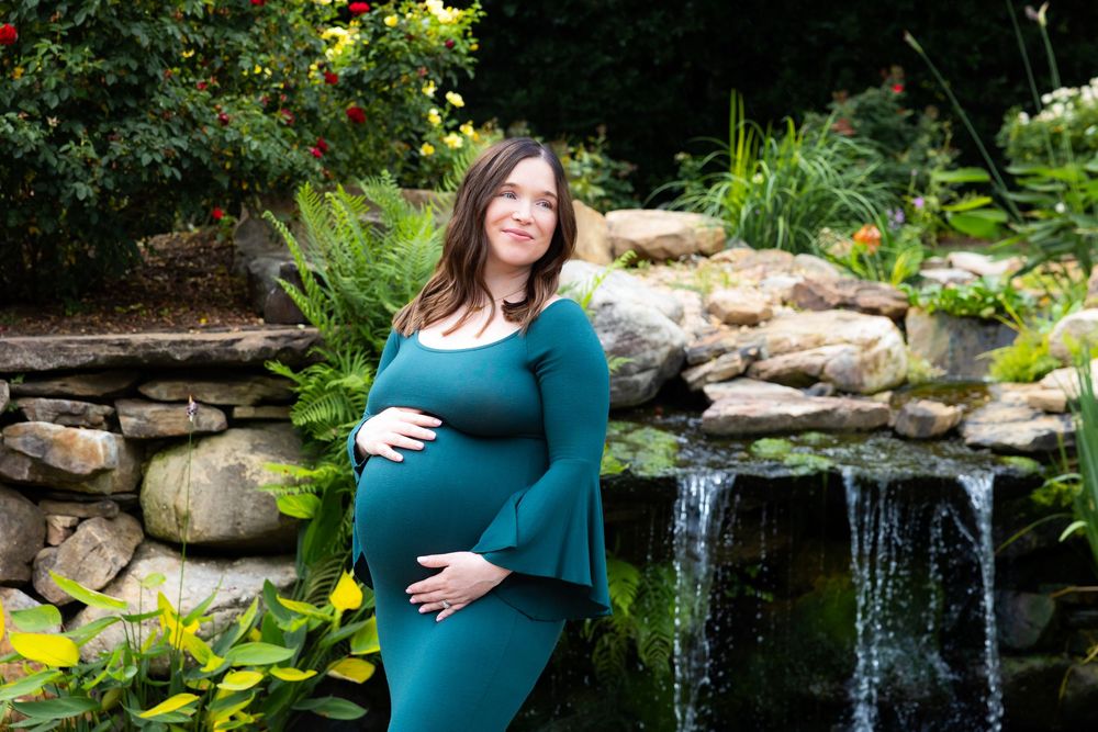Expectant mother in teal dress poses in front of garden waterfall and stone wall surrounded by lush greenery.