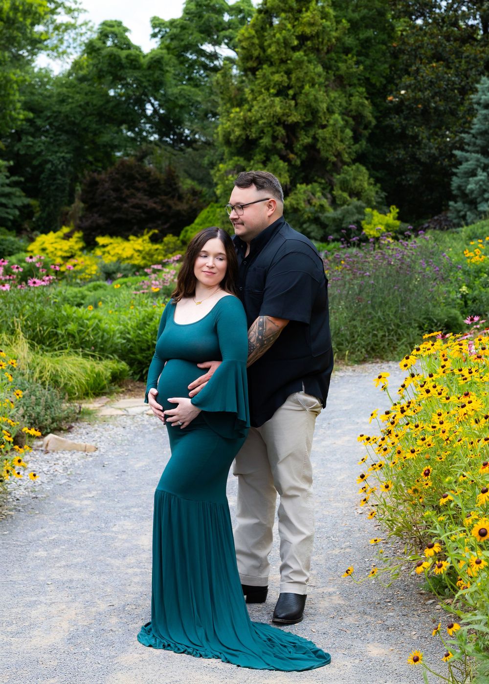 A couple shares an intimate moment on a garden path lined with vibrant yellow wildflowers and greenery during a maternity photoshoot