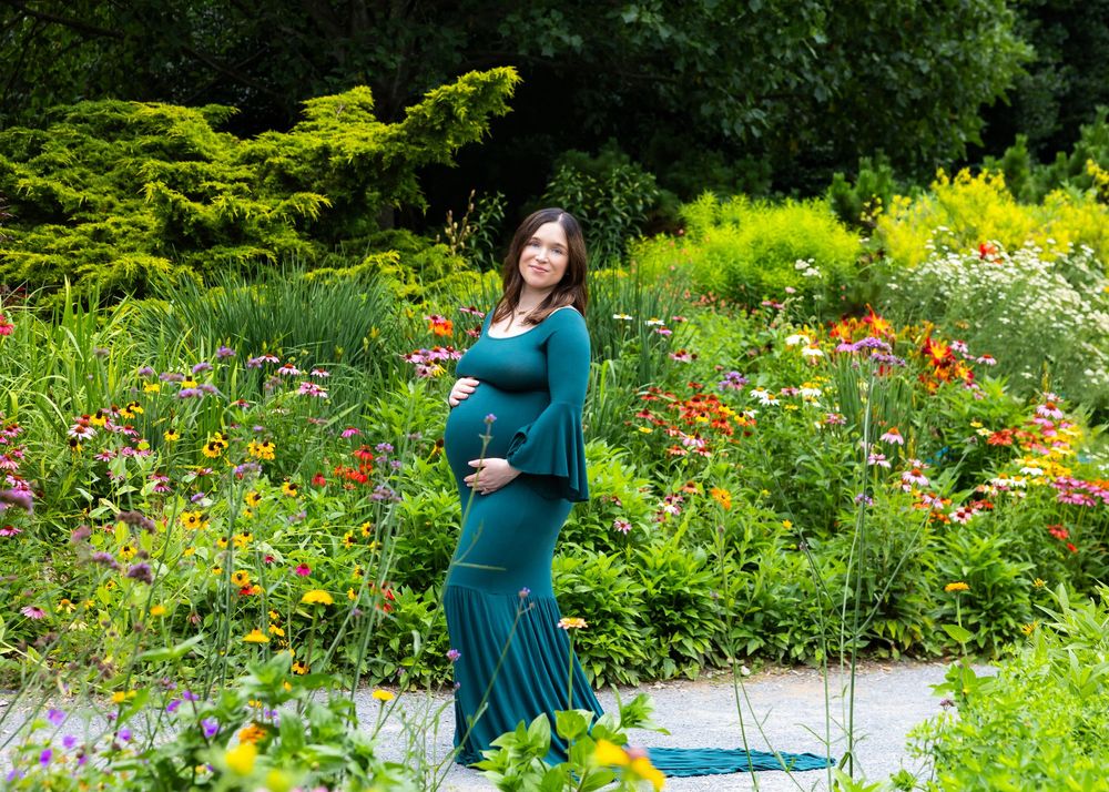 Maternity photoshoot in a teal dress stands among vibrant wildflowers in a lush summer garden setting.