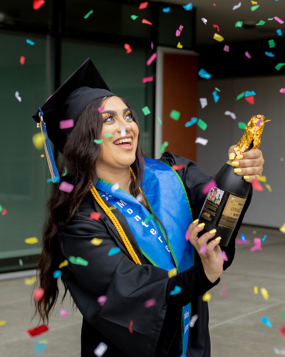 Graduate celebrates with colorful confetti falling while holding diploma and wearing traditional cap and gown.