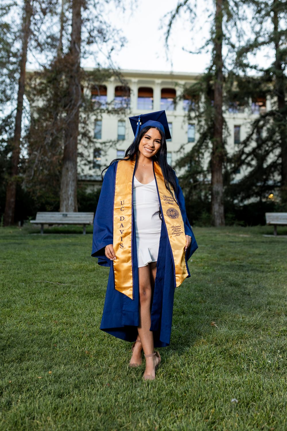 Graduate in blue and gold regalia poses on grassy lawn during outdoor commencement photo session.