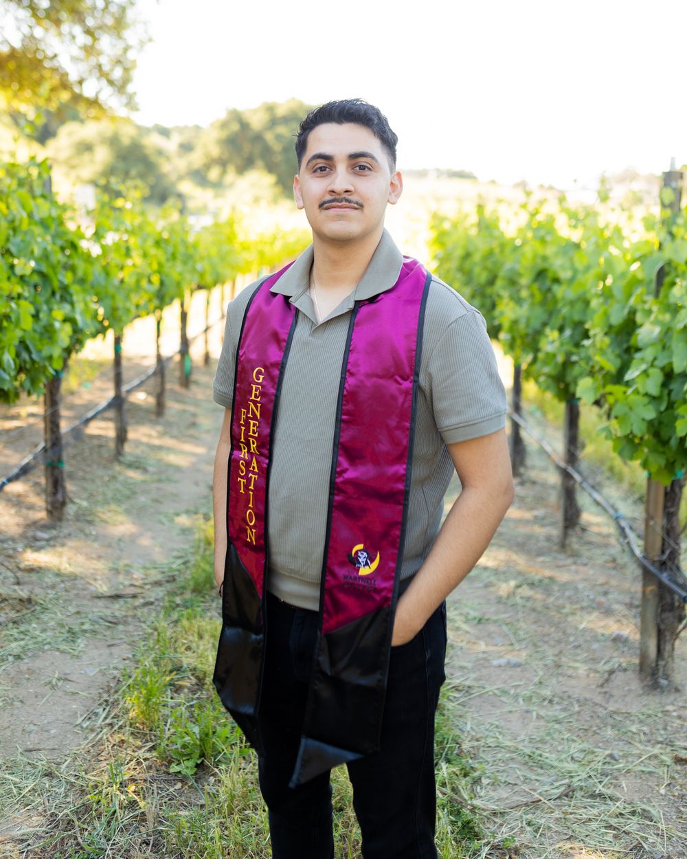 Person wearing a magenta vest poses in a sunny vineyard with rows of grapevines in the background.