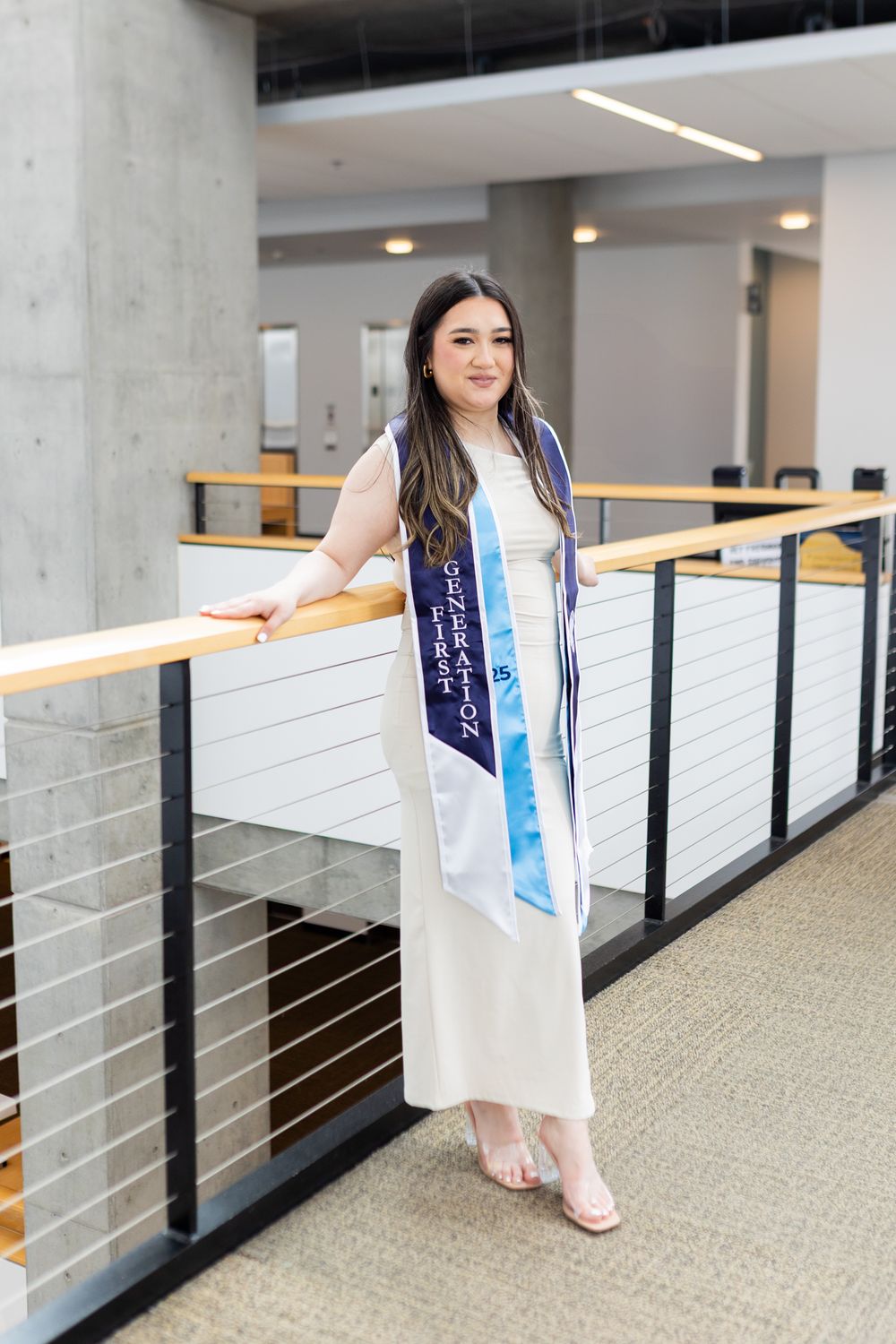 Graduate in white dress and graduation stoles stands in modern building hallway with concrete pillars and metal railings.