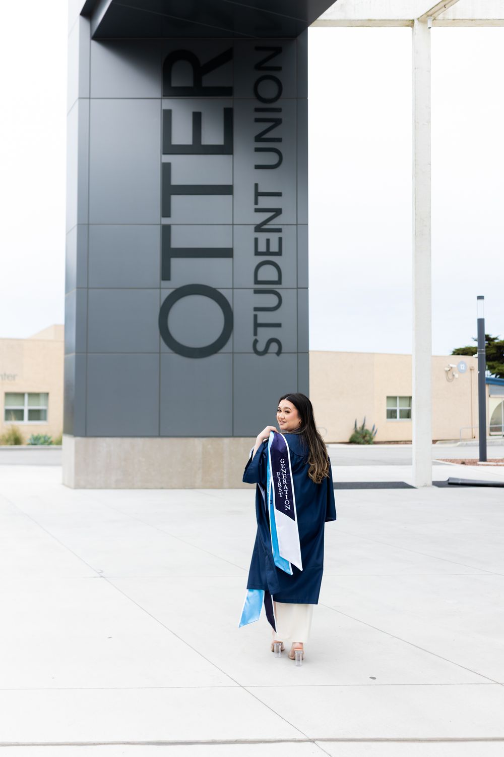 Graduate in blue cap and gown poses in front of Otter Student Union building sign on graduation day.