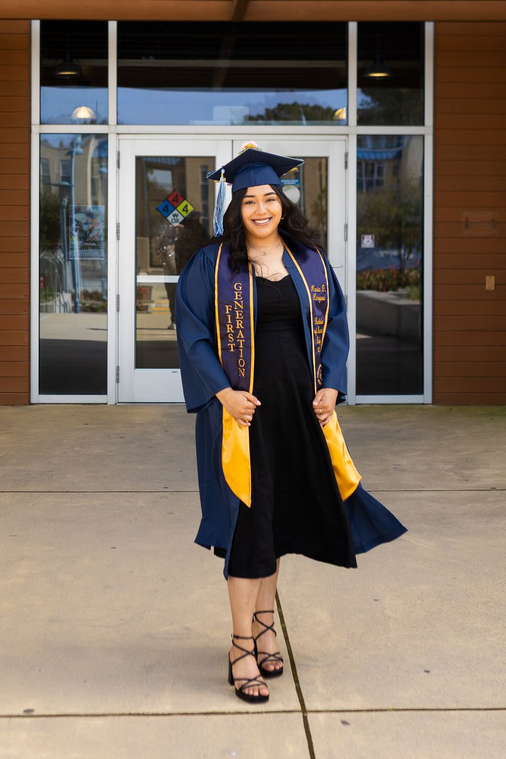 Graduate wearing navy blue cap and gown with gold stole stands proudly outside building entrance.