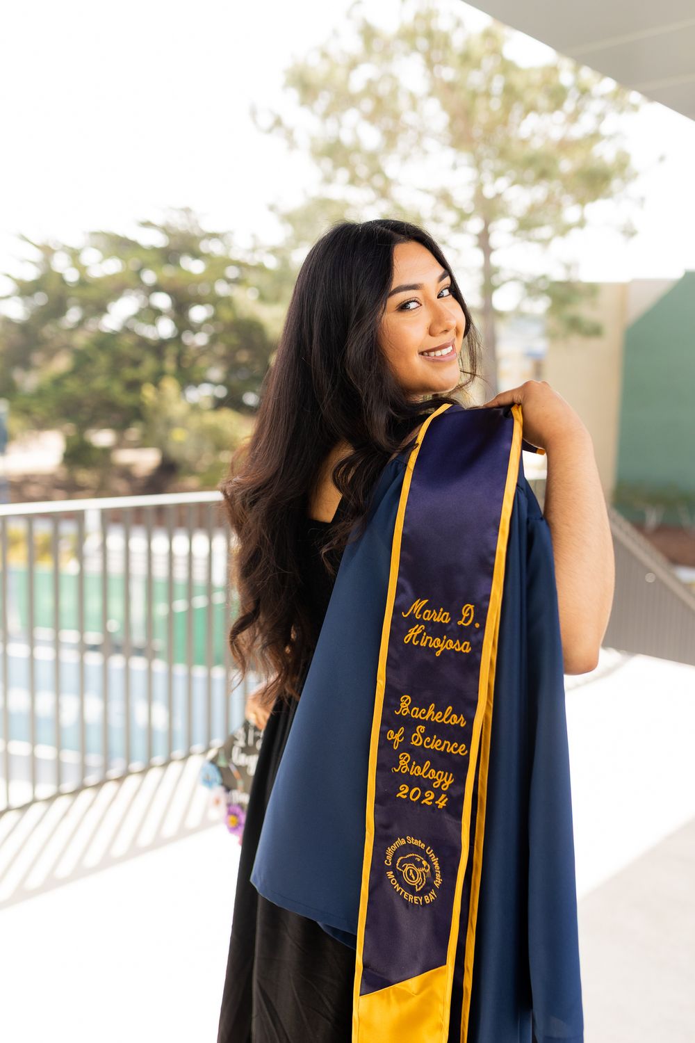 Graduate in navy blue academic regalia with gold stole poses outdoors during graduation photo session.