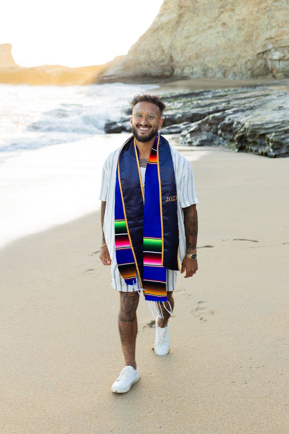 A person wearing a colorful graduation stole walks along a sandy beach near rocky cliffs at sunset.