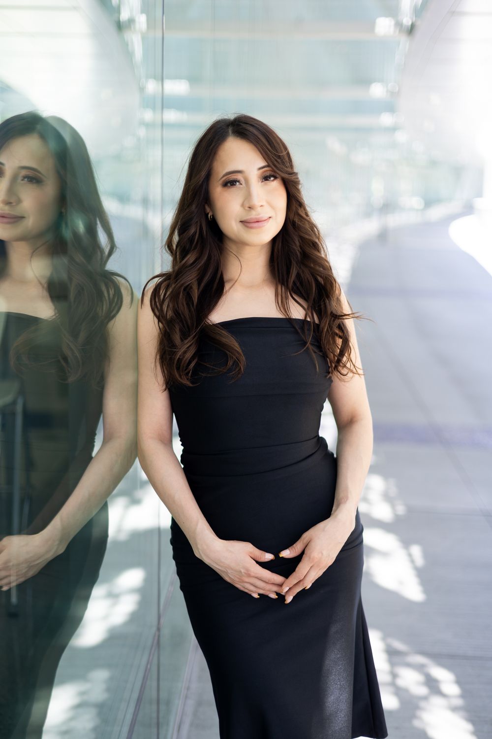 A woman in a black dress poses elegantly against a reflective glass wall with multiple mirror reflections.