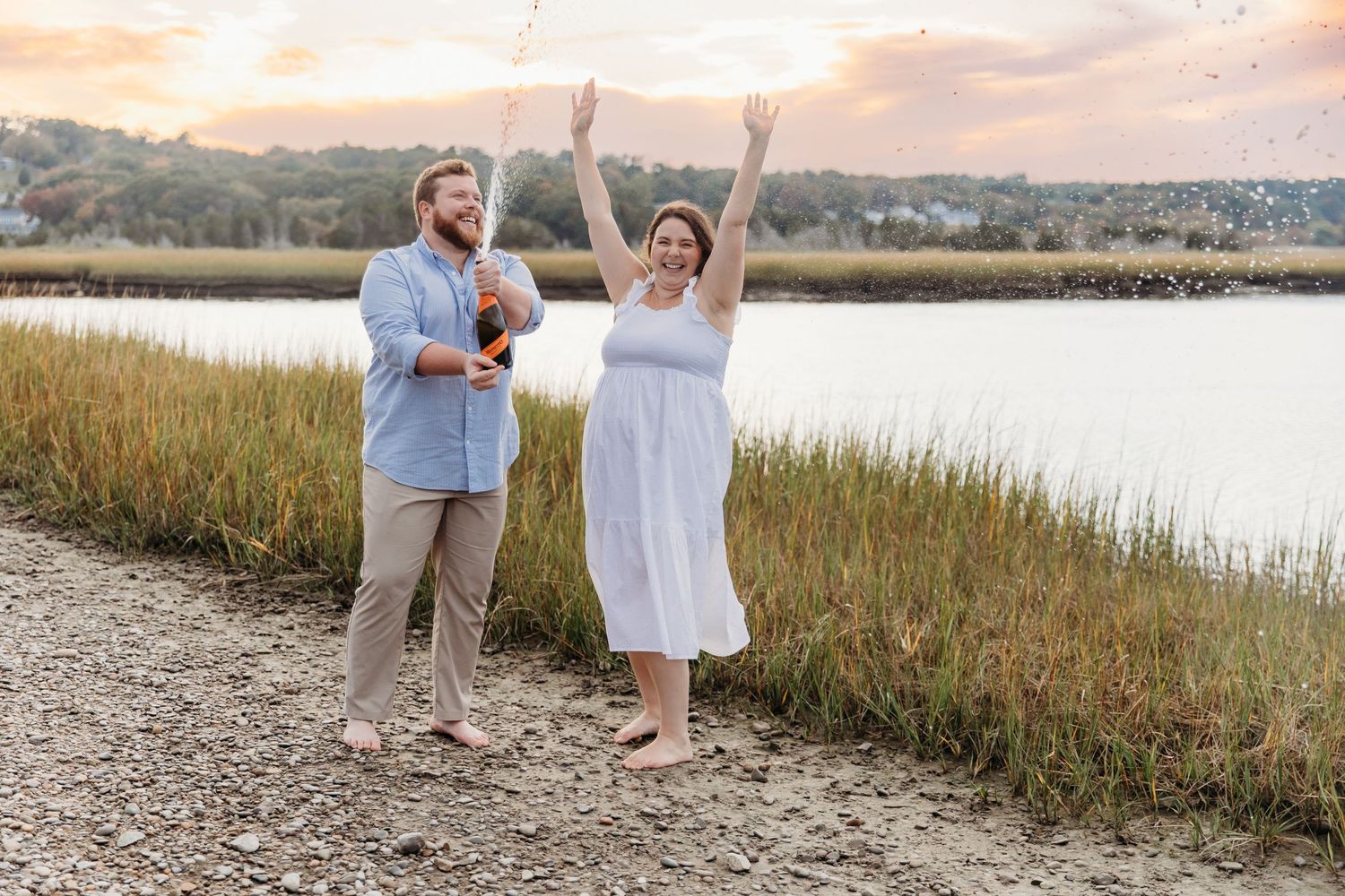 Two people celebrating joyfully at sunset on a coastal path beside marsh grass and water.