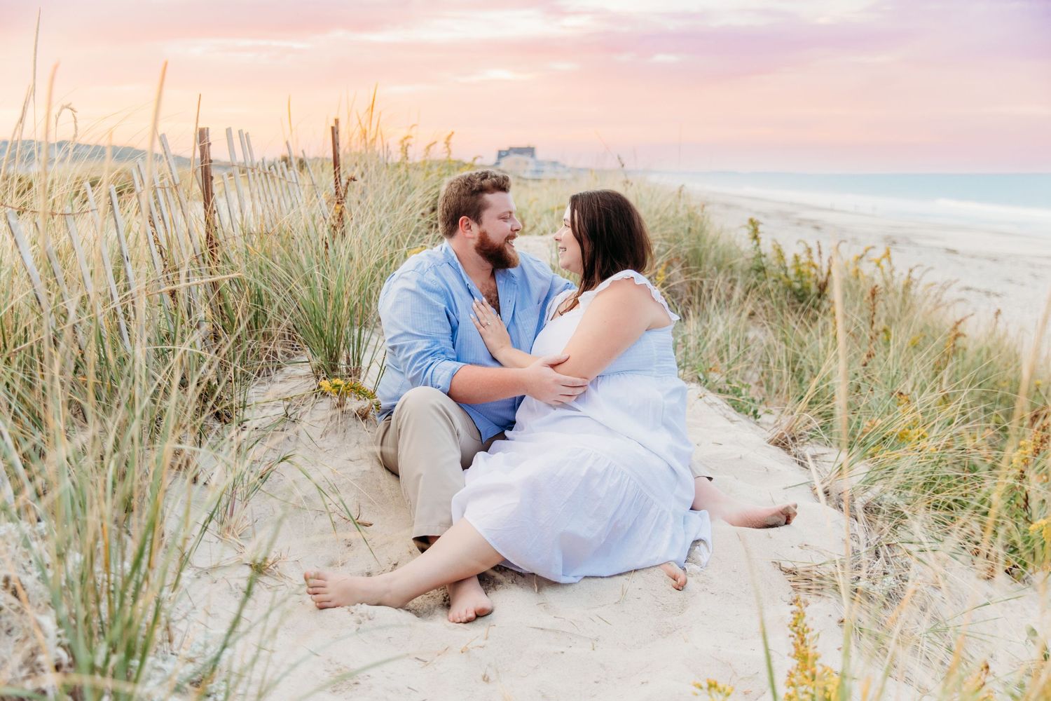 A romantic couple sits together on a sandy beach dune at sunset with sea grass swaying in the breeze.