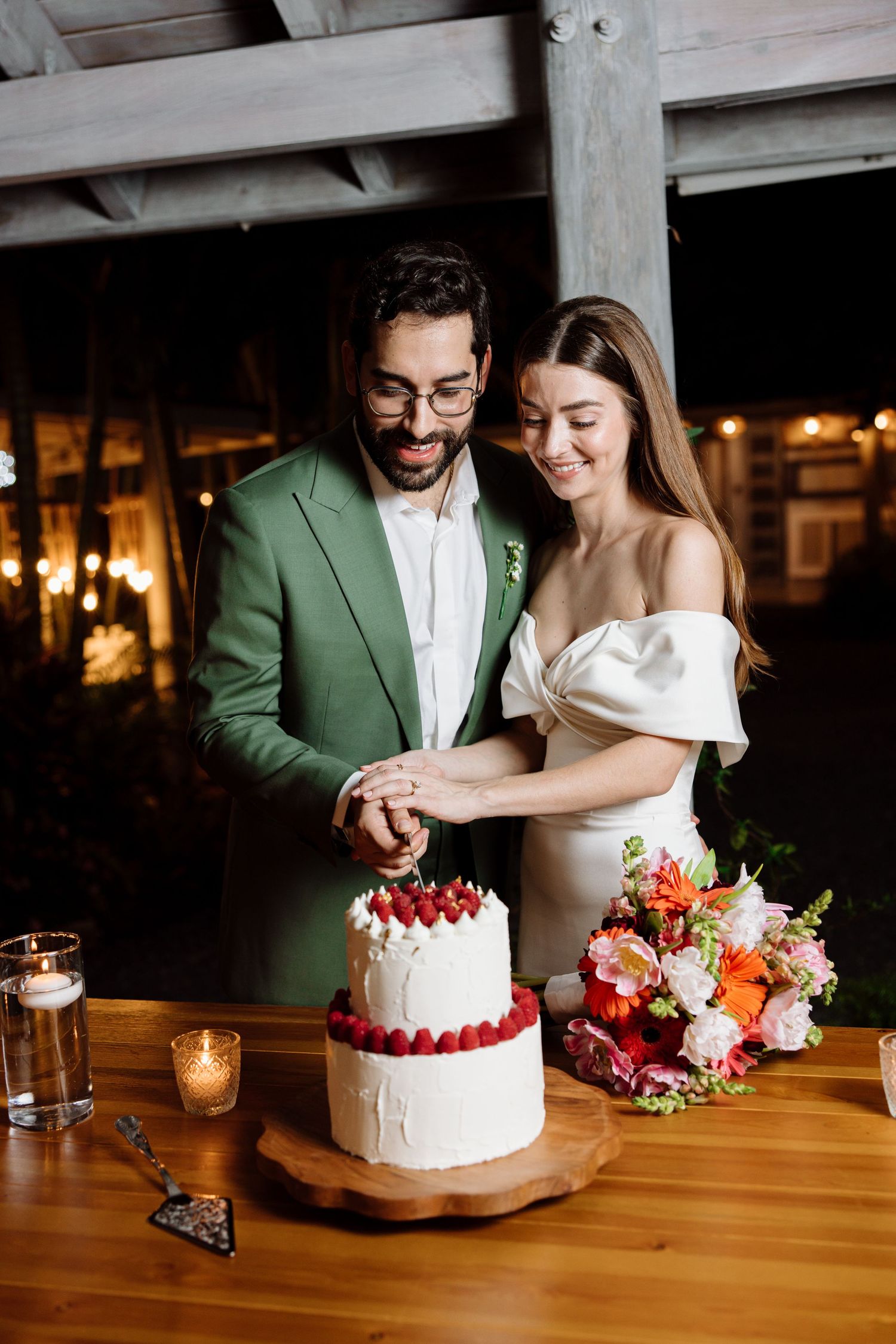 Couple cuts their two-tier white wedding cake decorated with red berries while surrounded by candles and floral decor.