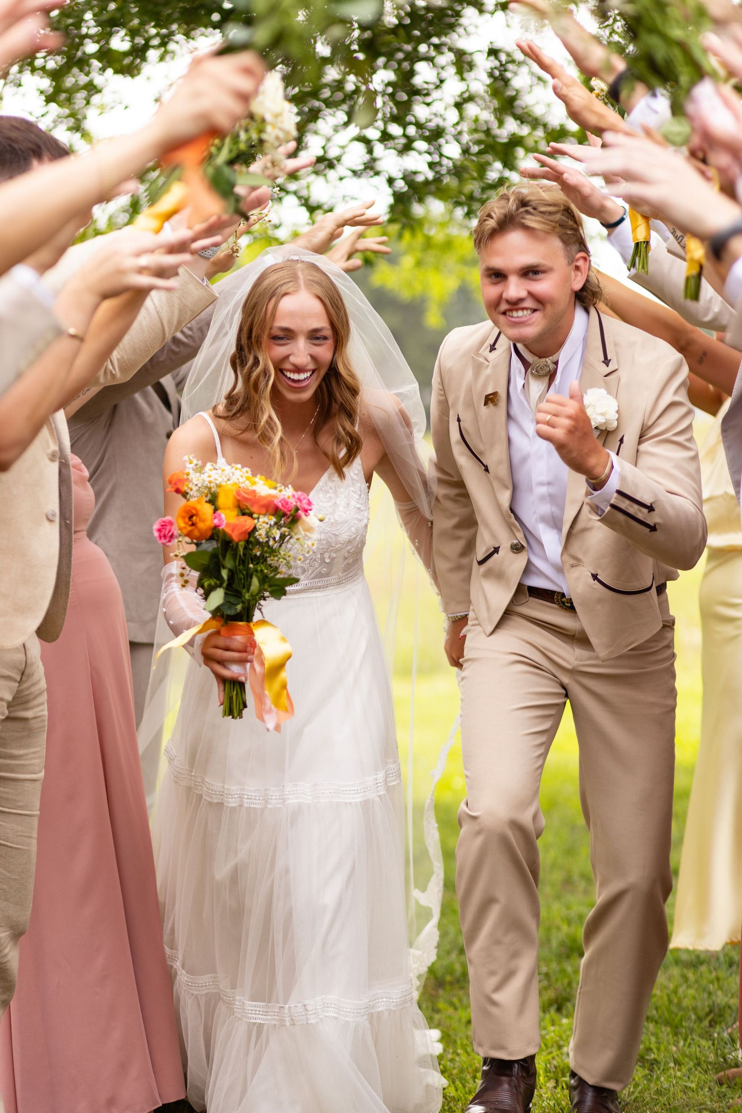 Newlyweds walk through tunnel of guests tossing flower petals while bride holds vibrant orange bouquet at outdoor celebration.