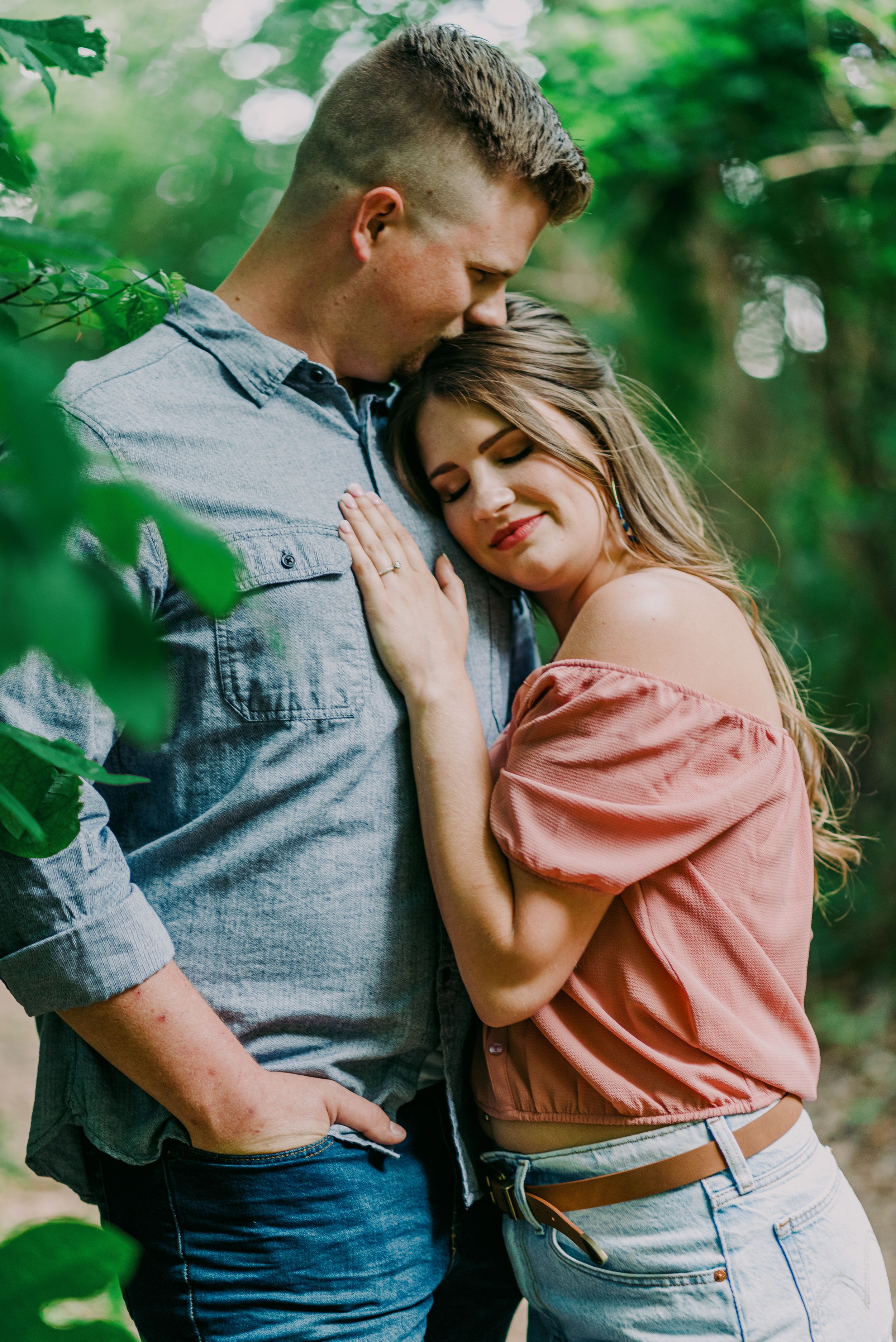 Couple sharing a tender embrace surrounded by lush green leaves during outdoor engagement session.